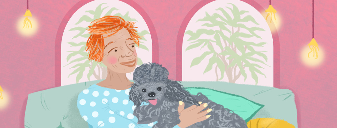 A person with red hair smiles at a fluffy gray poodle. A couch and soft lights float behind her.