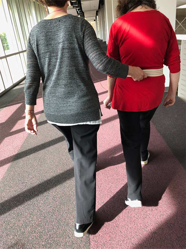 Two women walk side by side, one gently guiding the other with a waist belt.