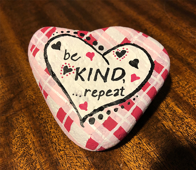 A heart-shaped rock is painted in pink and white plaid with the words be kind, repeat set into a smaller white heart.