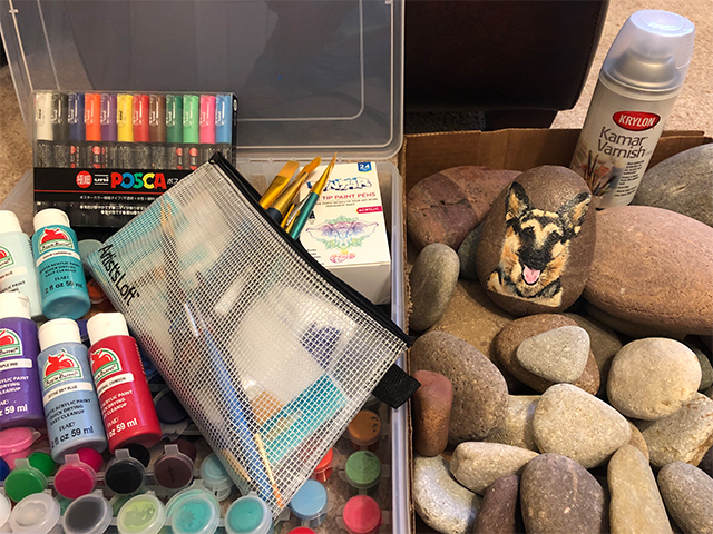 Project supplies including craft paints, brushes, and smooth rocks. One rock is painted with a realistic portrait of a german shepherd.