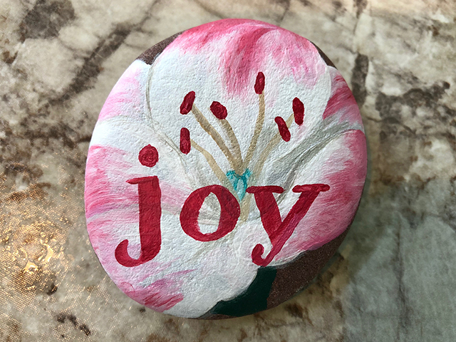 A rock is painted with the word joy over a pink tropical flower.
