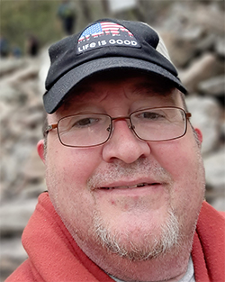 Dan Glass in a hoodie and ball cap, smiling in front of a natural landscape.