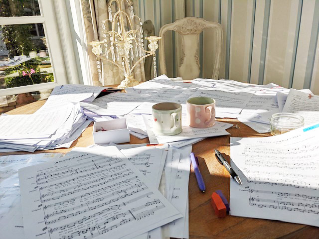 Sheet music papers, pens, and mugs spread out on a sunny table.