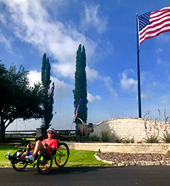 Person on a recumbent bike in front of an American flag and a statue of a long-horn bull.